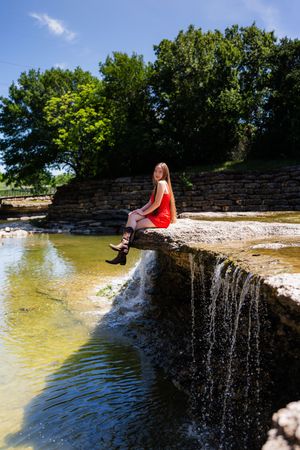 Woman in red dress and black boots sitting on stone ledge over waterfall in sunny park. Senior session Airfield Falls Fort Worth Texas