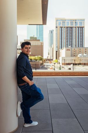 Young man in dark shirt and jeans leaning against a column on an urban rooftop terrace with city skyline backdrop. Senior session FTW