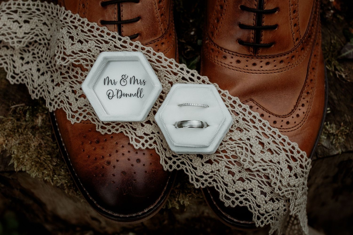 Wedding rings and hexagonal name tag displayed on vintage lace next to brown leather shoe.