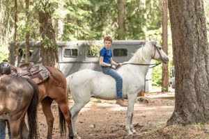 A rider on a white horse stands near a brown horse in a wooded camping area with an RV trailer visible.