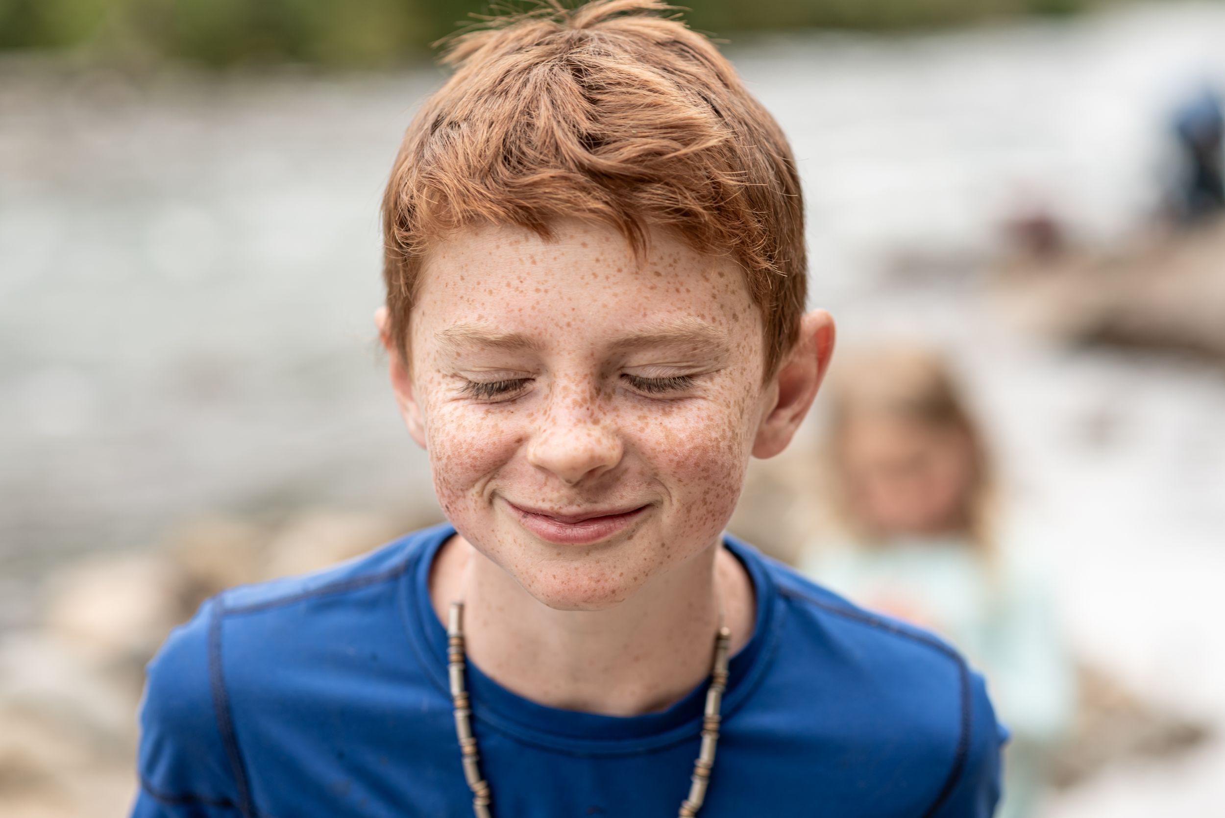 A youth in a blue shirt smiles peacefully with eyes closed outdoors.