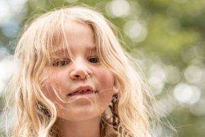 A child with flowing blonde hair smiles against a soft bokeh background of green foliage.