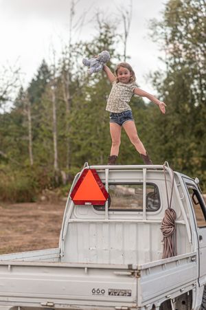 A young person jumps joyfully from the roof of a white pickup truck with a red safety triangle against a forest backdrop.