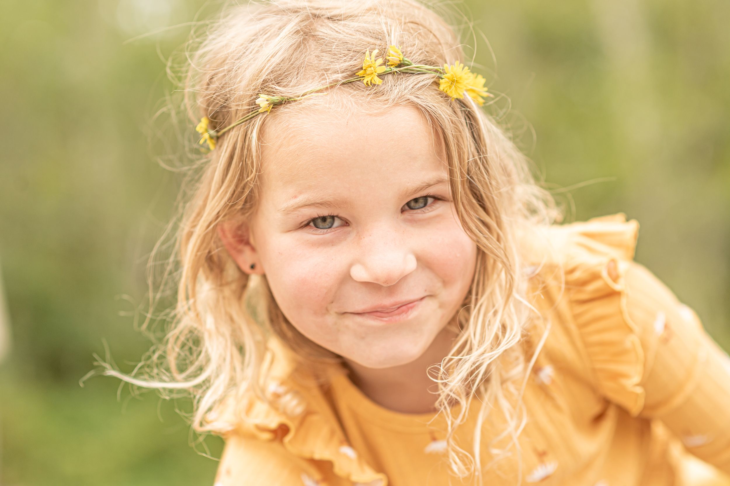 Child wearing yellow dress and floral crown smiles in soft sunlit outdoor portrait.