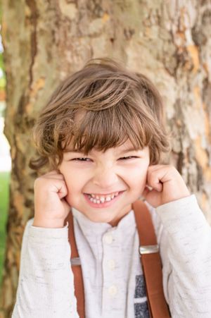 Young person in sweater and suspenders poses playfully by tree trunk.