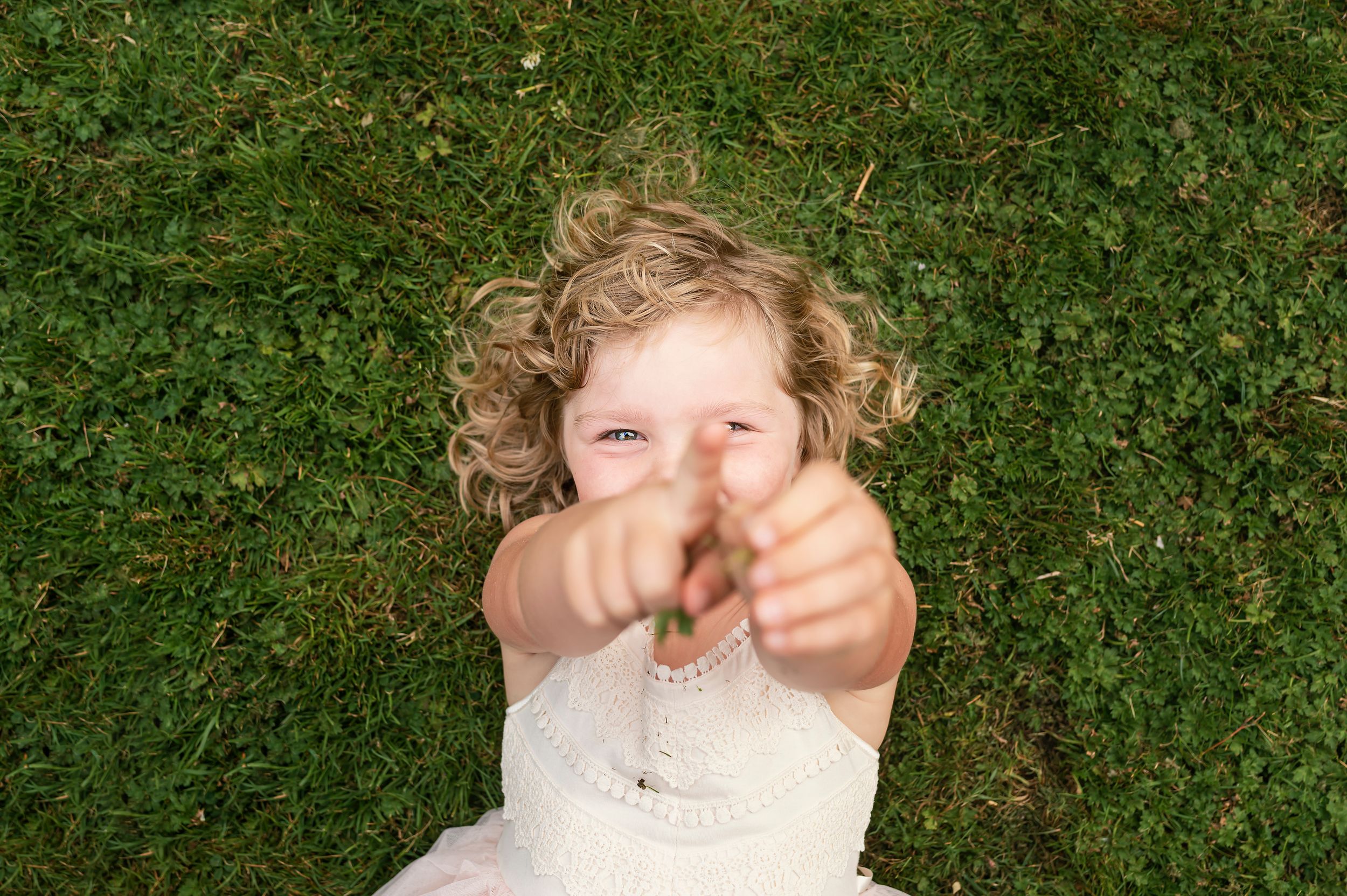 Child lying on grass points fingers playfully at camera.