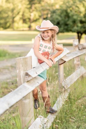 Child in cowboy boots and hat leans against rustic wooden fence in countryside setting.