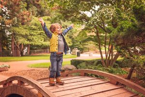 Child in yellow jacket stands triumphantly with arms raised on wooden bridge.