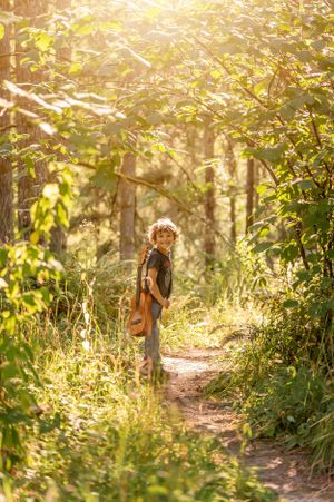 Child carrying guitar stands on sunlit forest trail surrounded by lush greenery.
