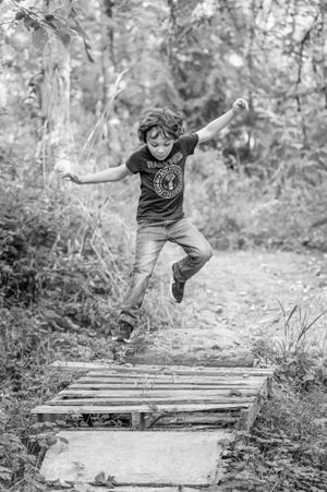 Child leaps joyfully over wooden bridge on forest trail in black and white photo.