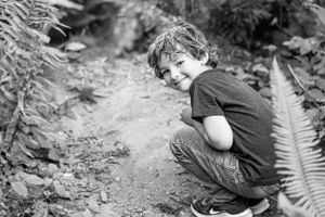 Black and white photo of someone crouching on a forest trail surrounded by ferns.