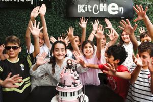 Children celebrating with raised hands around a piano-themed birthday cake at a party.