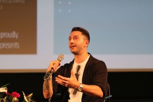 A speaker wearing a dark blazer and white t-shirt gestures while holding a microphone during a presentation.