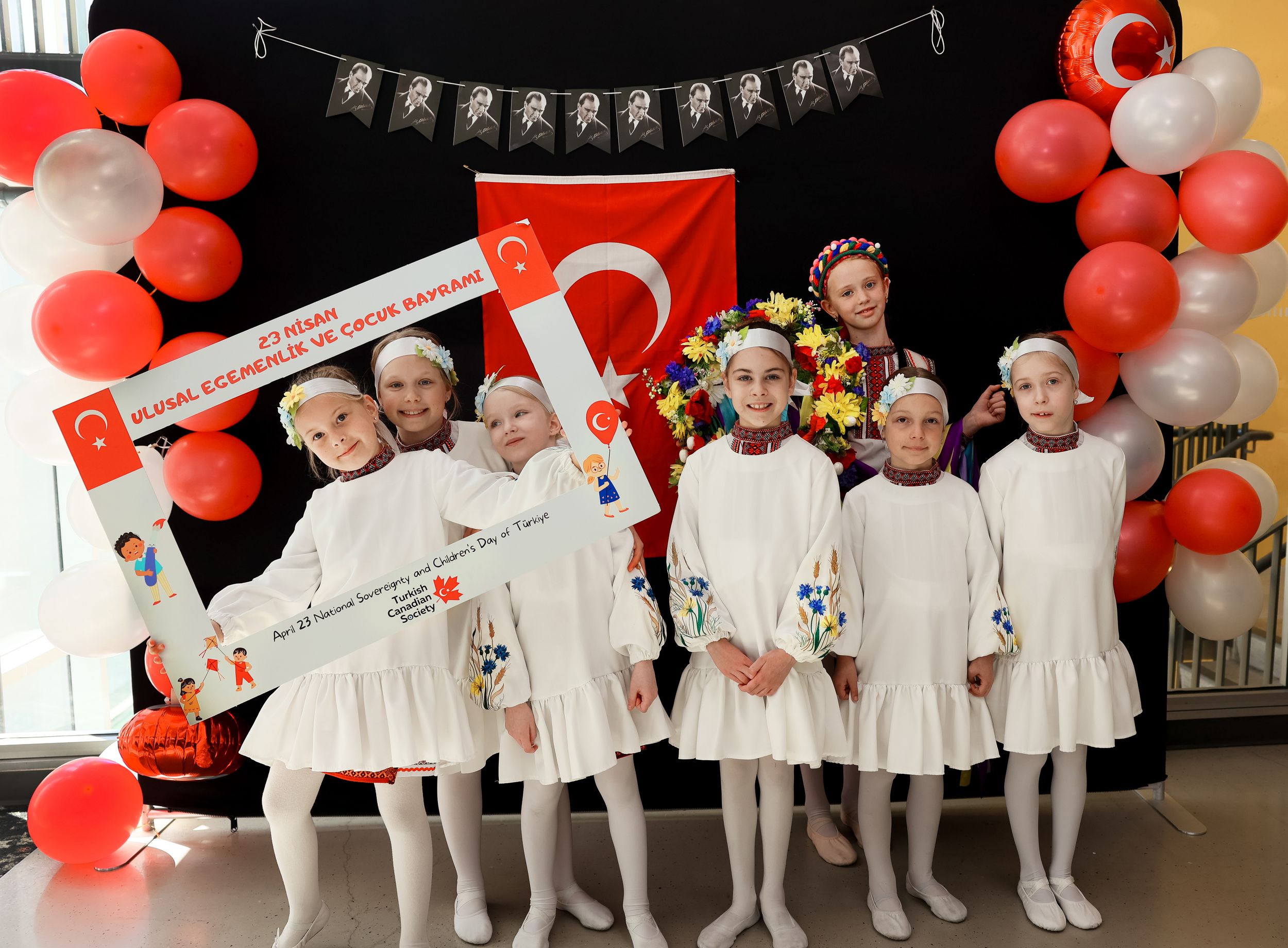 A group in white outfits pose at a celebration decorated with red and white balloons and Turkish flag.