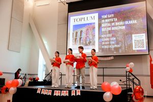 Performers in white pants and red shirts stand on stage with balloons and a presentation screen during an event.