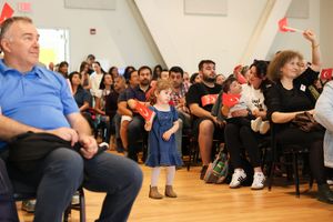 Crowd watches intently during indoor event as small child in blue dress stands in front of seated audience.