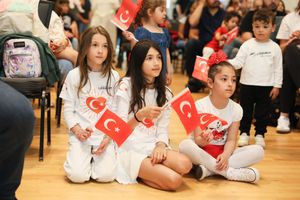 Students wearing white outfits hold Turkish flags while sitting on a wooden floor during a cultural celebration.