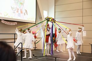 Young ballet dancers in white costumes performing a traditional maypole dance with colorful ribbons on stage.