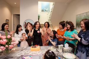 A joyful group gathers around a table with pink flowers and cake at what appears to be a celebration or party.