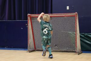 A young athlete in a green uniform stands in front of a hockey goal net in an indoor sports facility.