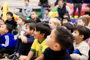 Children sit together on the floor watching and reacting excitedly at a sporting event or assembly.
