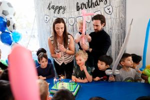 A birthday celebration with guests gathered around a cake with candles while blue and pink balloons decorate the party space.