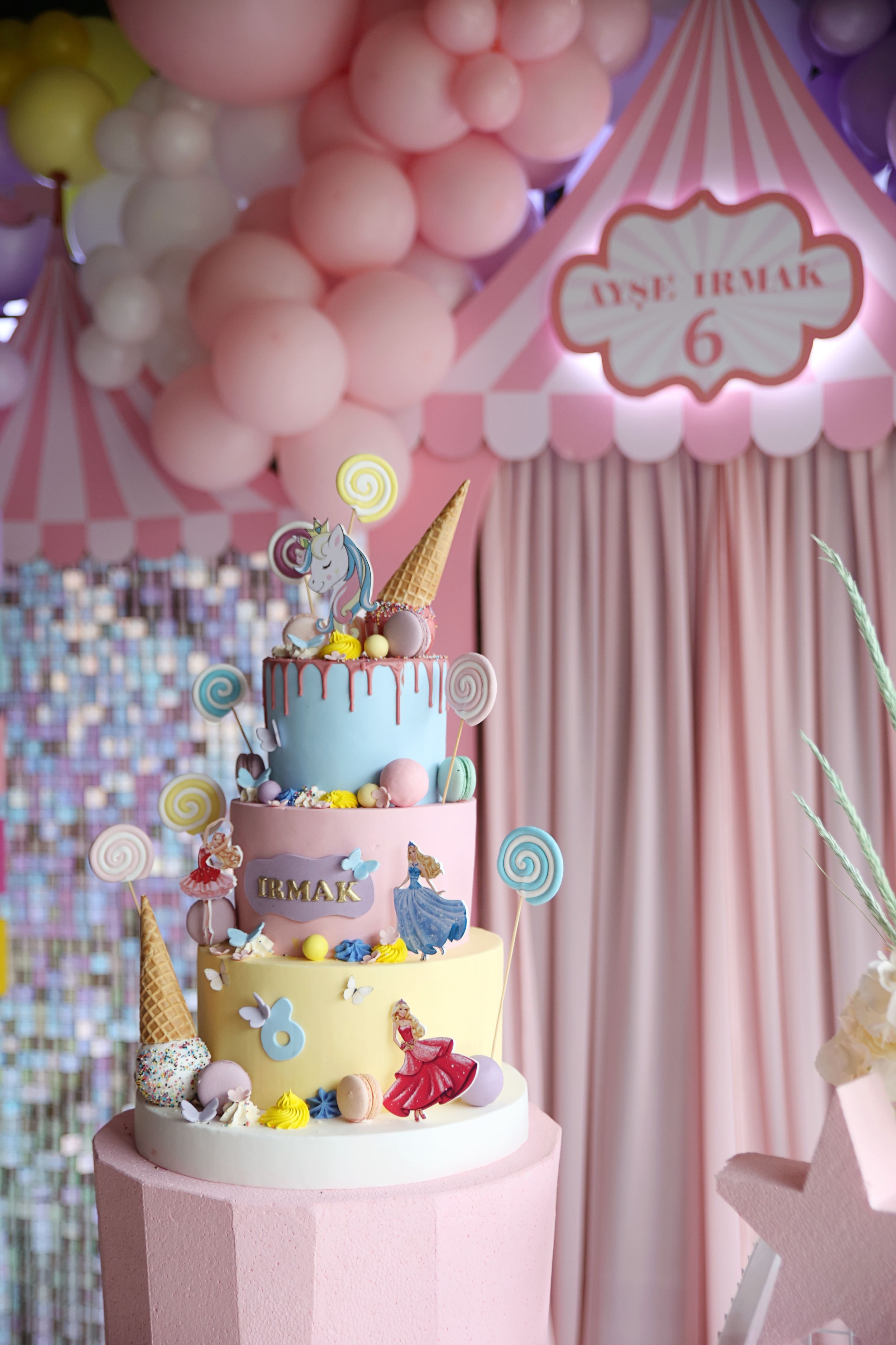 Whimsical three-tiered birthday cake decorated with lollipops and candy on a pink stand against a balloon backdrop.