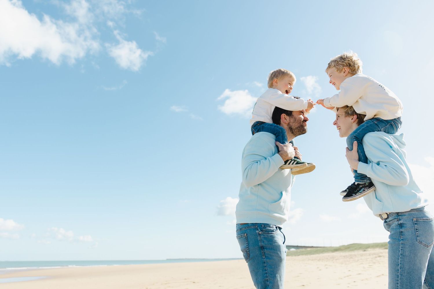 A couple stands on a sunny beach lifting young children on their shoulders against a blue sky backdrop.