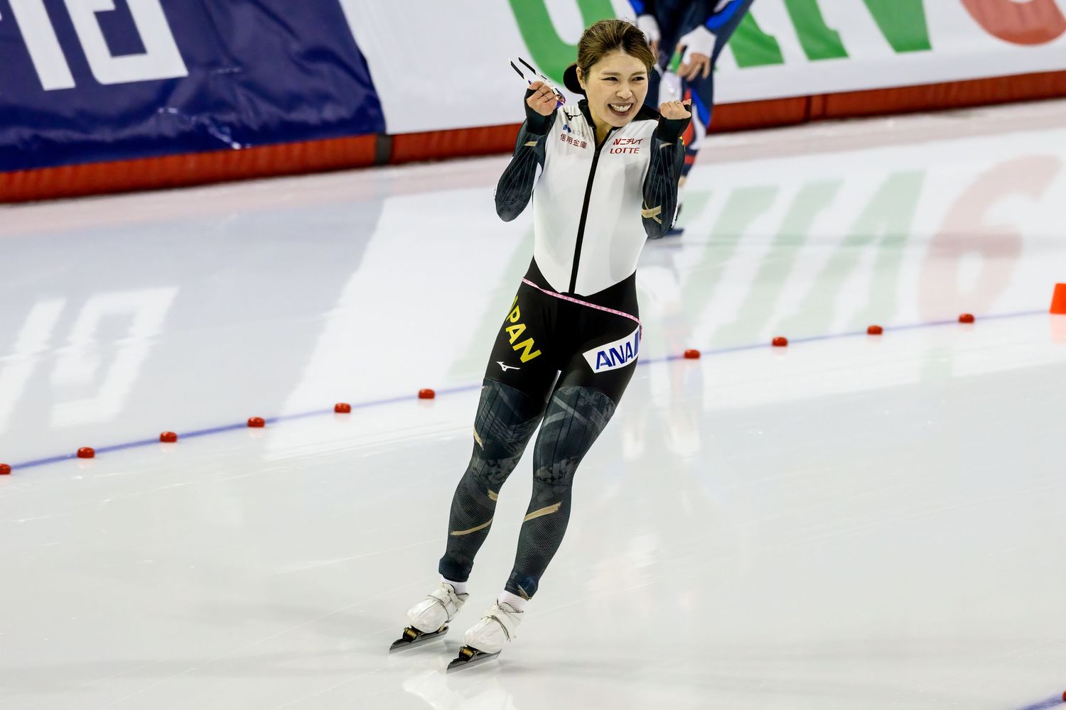 Speed skater glides across ice rink with arms raised in celebration while wearing dark uniform with green accents.