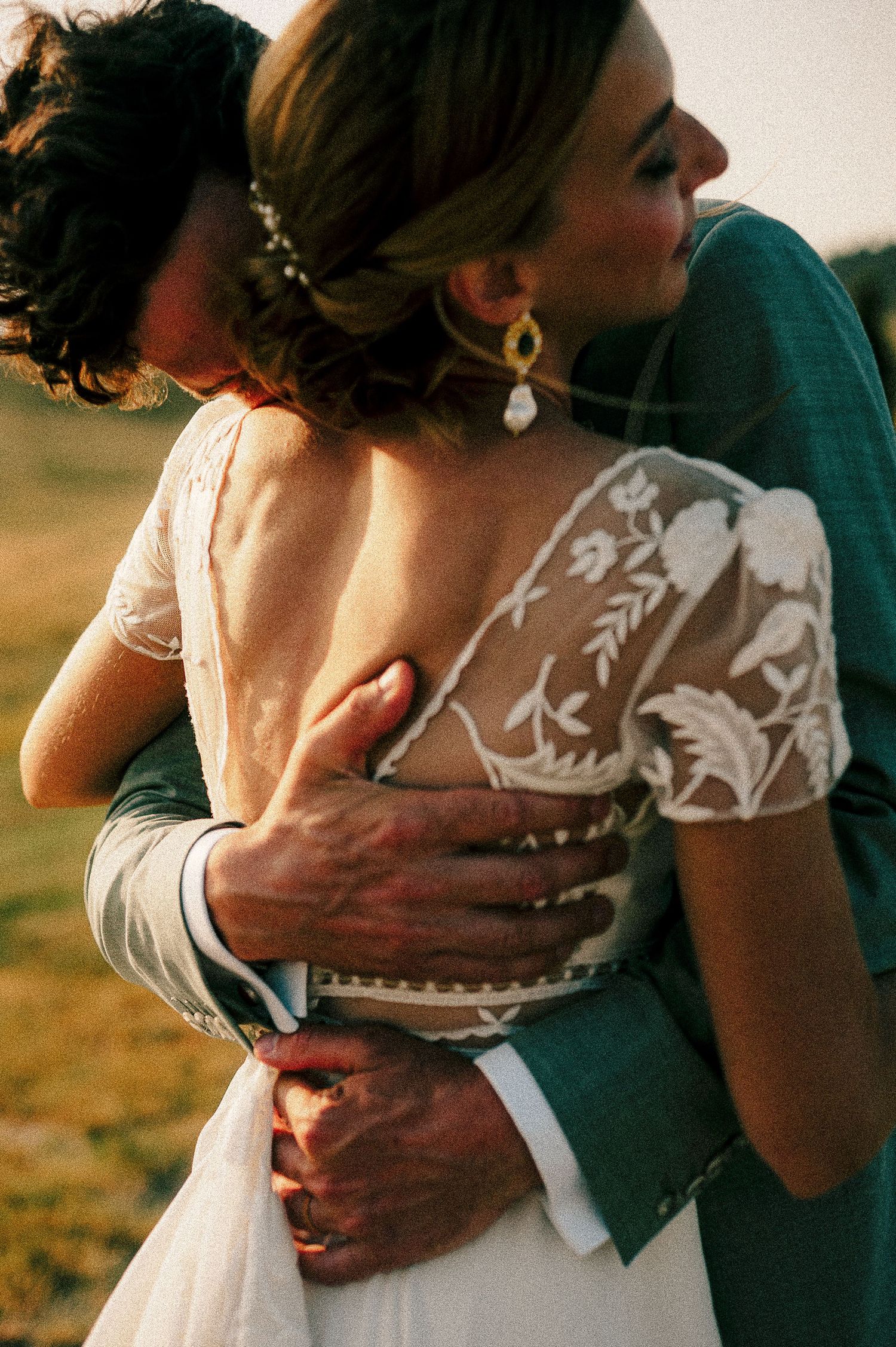 Intimate embrace between newlyweds showing detailed lace wedding dress and green suit during outdoor celebration.