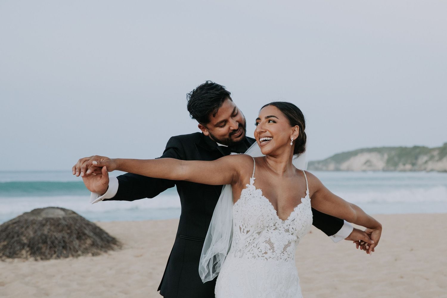 Joyful newlyweds dancing together on a scenic beach with cliffs in the background.