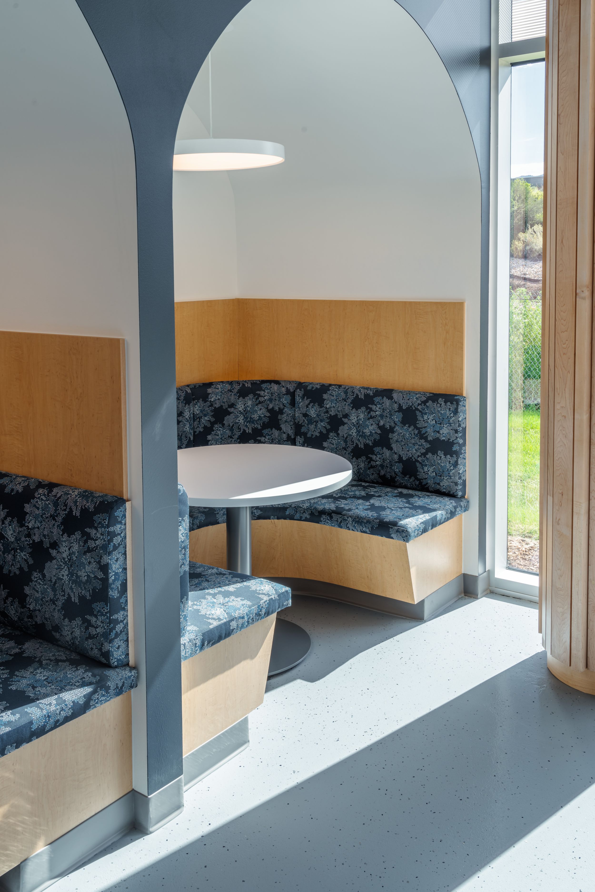 Modern booth seating area with blue velvet upholstery, wooden panels, and arched divider in a bright space with window views.