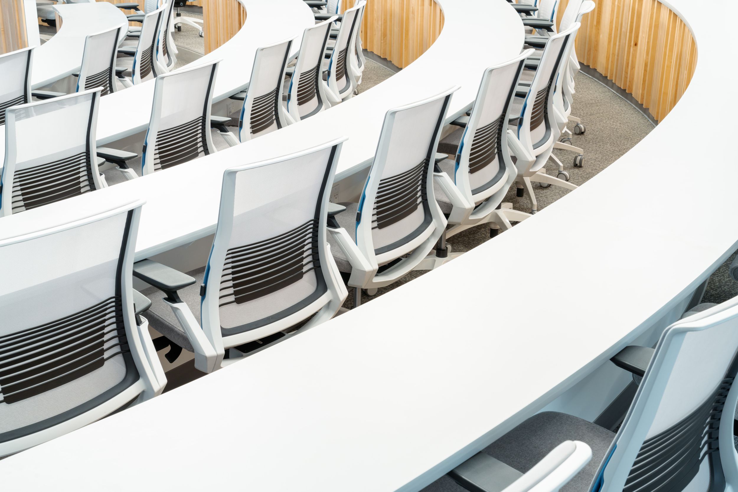 Curved rows of modern office chairs with grey mesh backs and wooden seats arranged in a lecture hall.