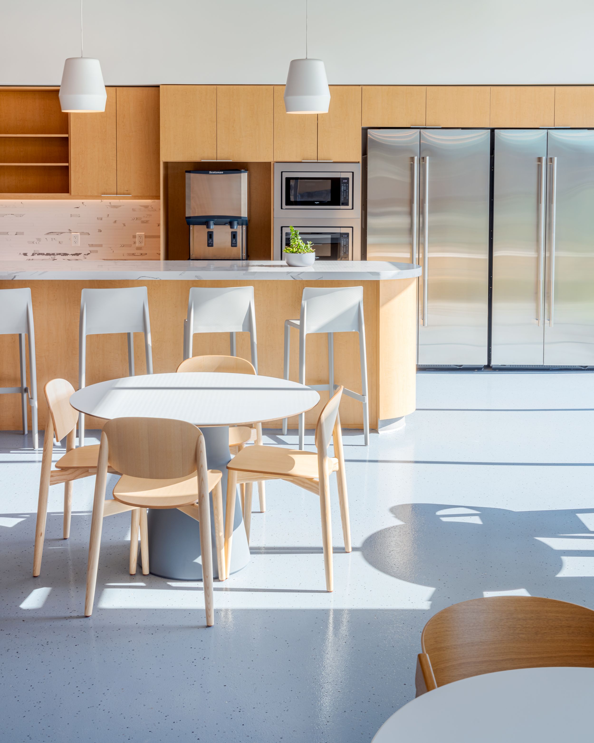 Modern office break room with light wood furniture, round white table, and stainless steel refrigerators in minimal design.
