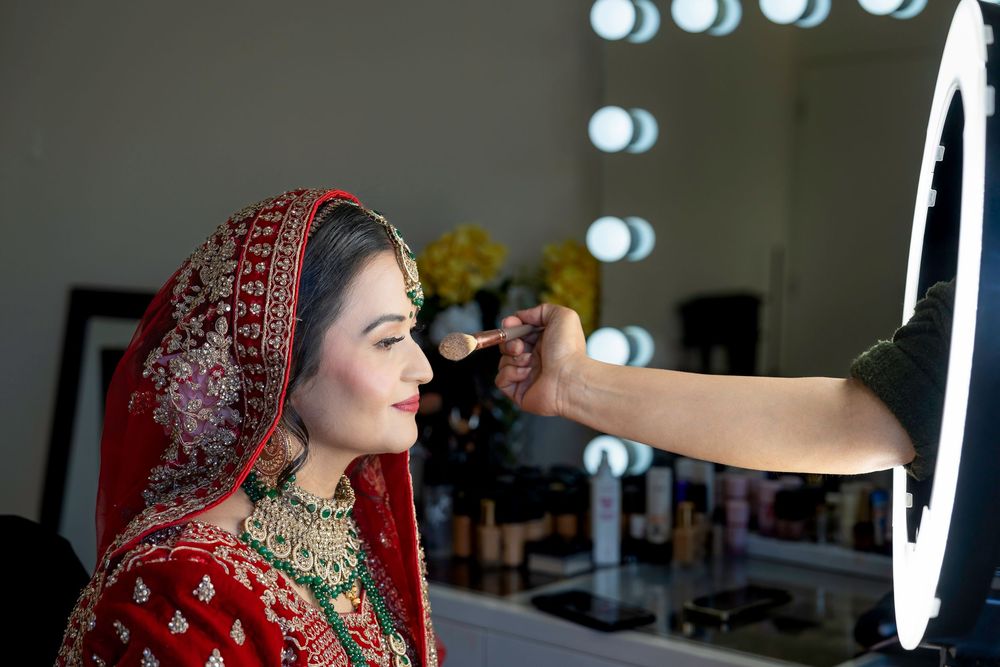A makeup artist applies cosmetics to a bride wearing a traditional red and gold embroidered dress and headpiece.
