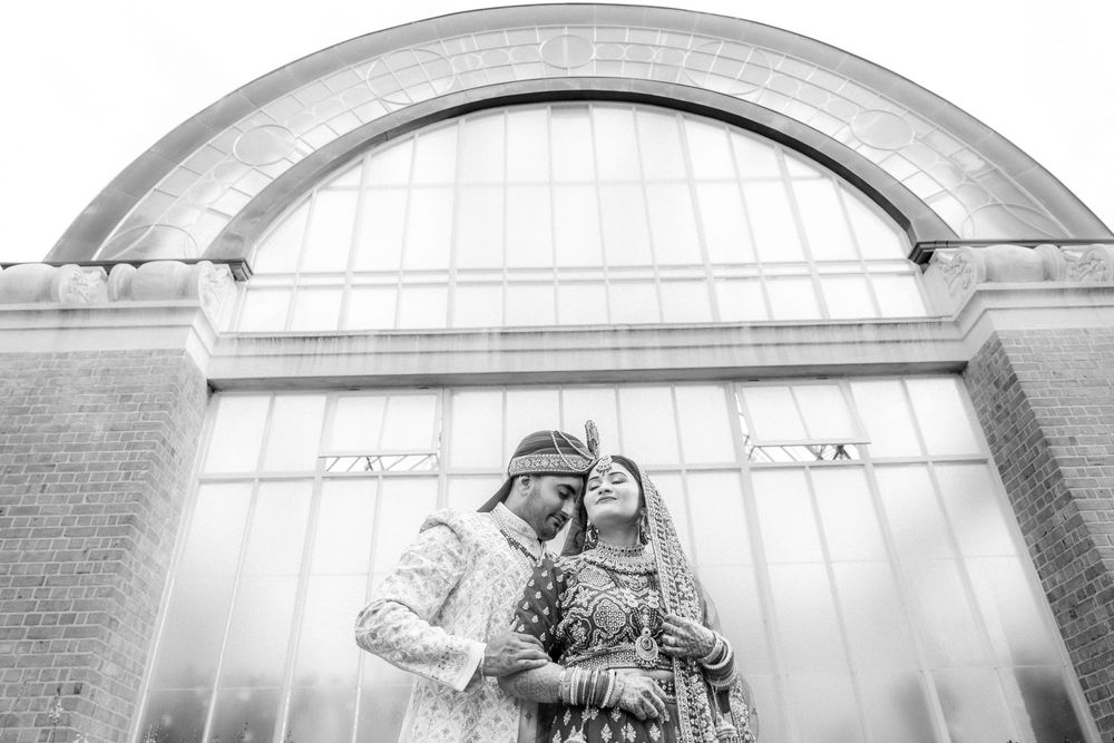 A romantic couple in winter attire embraces beneath an arched entryway in a black and white photograph.