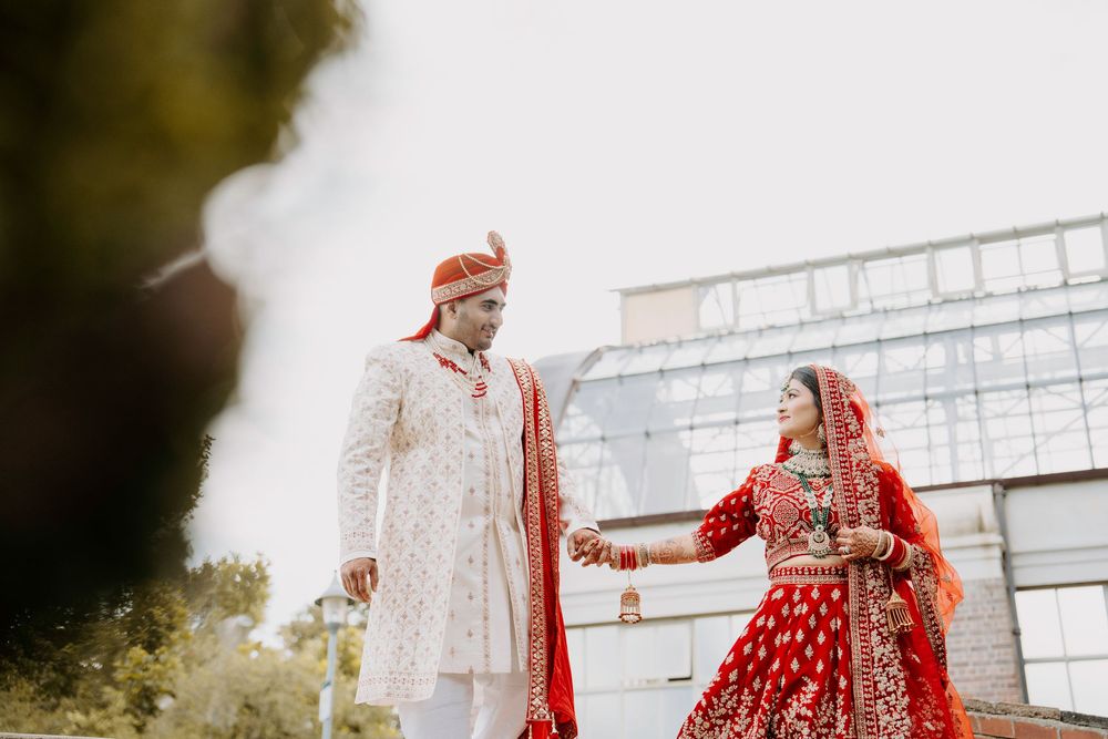 Traditional Indian bride and groom in red and ivory wedding attire hold hands against modern building backdrop.