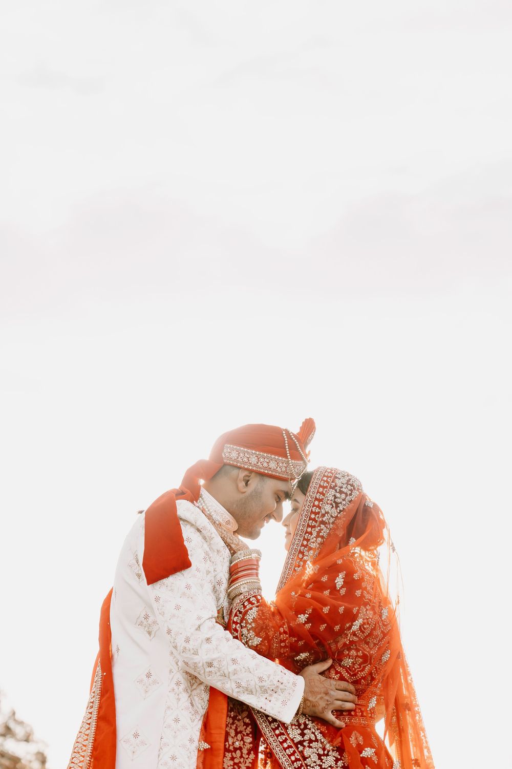 Two people in traditional red and white Indian wedding attire embrace under an overcast sky.