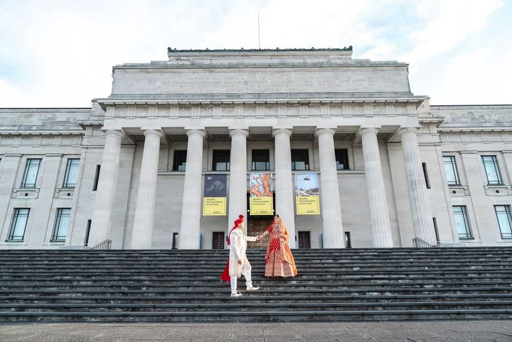 Two people in colorful clothing stand on the steps of a grand neoclassical building with white columns and facade.
