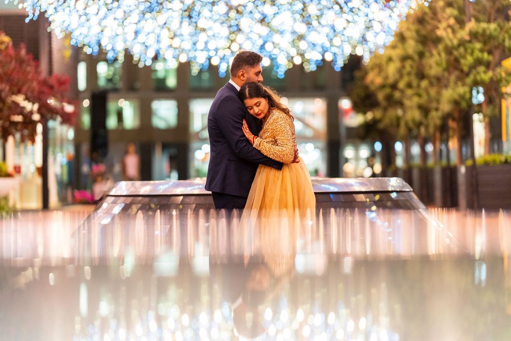 A romantic couple embraces near an illuminated fountain at dusk with twinkling lights overhead.