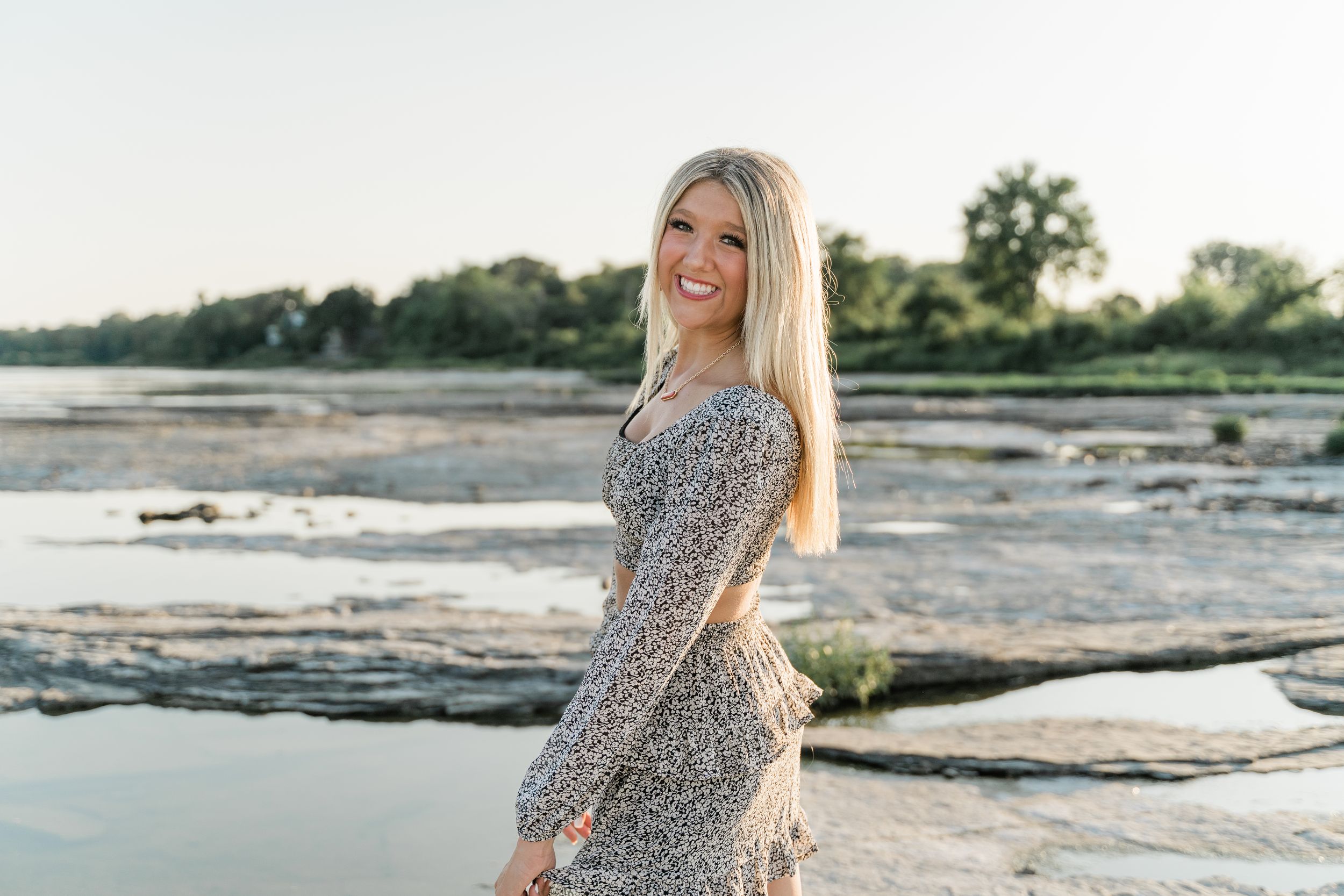 A person in a floral dress poses on rocky coastal outcroppings during sunset at the beach.