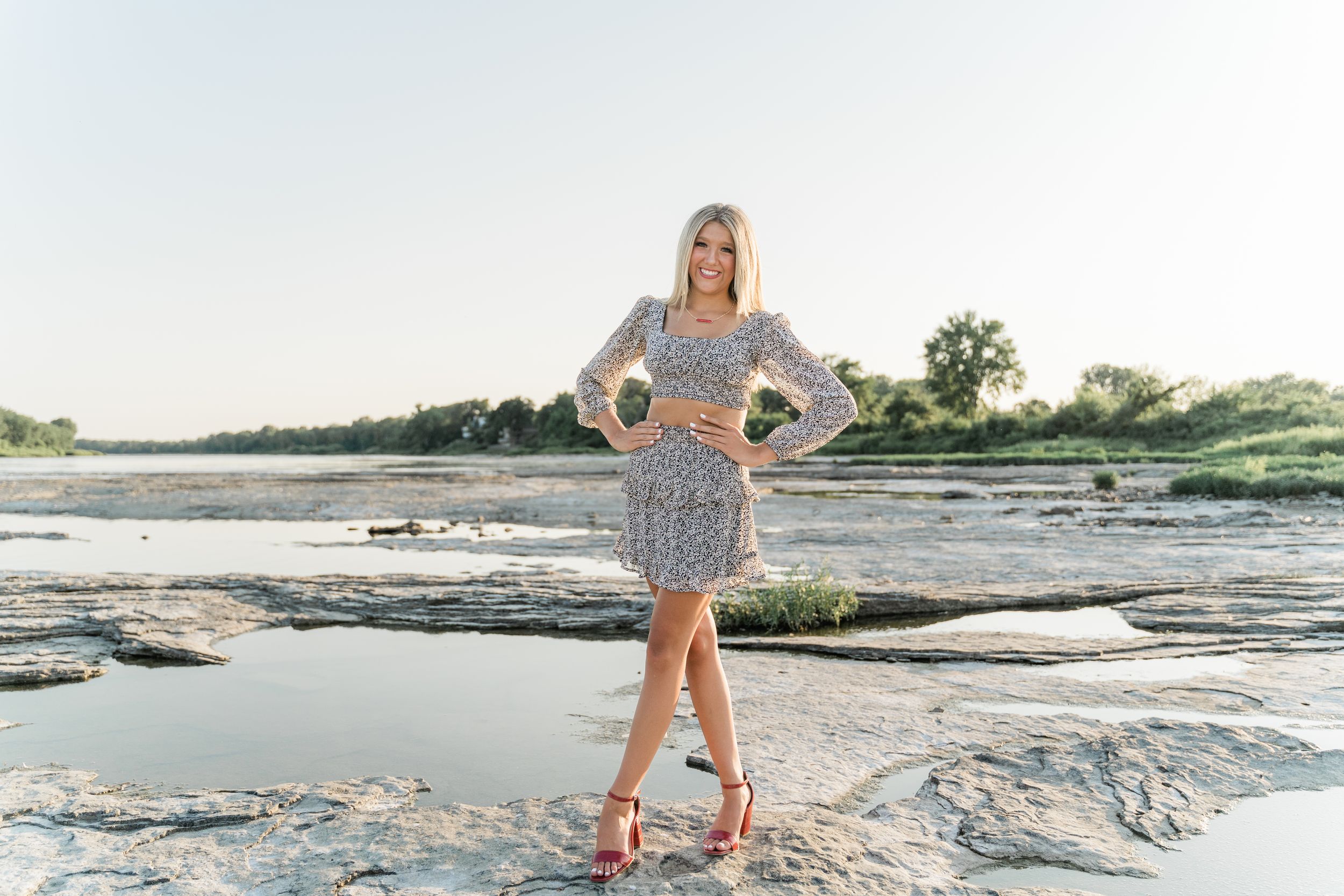 Woman in patterned dress poses on rocky riverbank during sunset with water and trees in background.