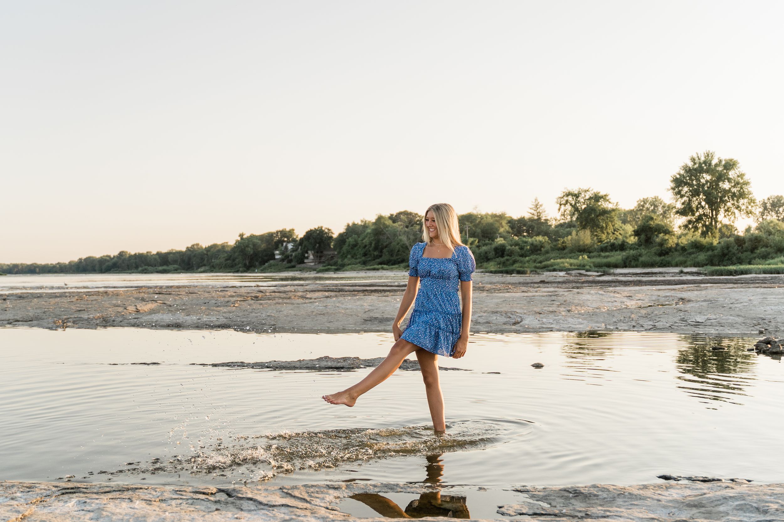 A person in a blue dress splashes through shallow water at sunset on a serene beach.