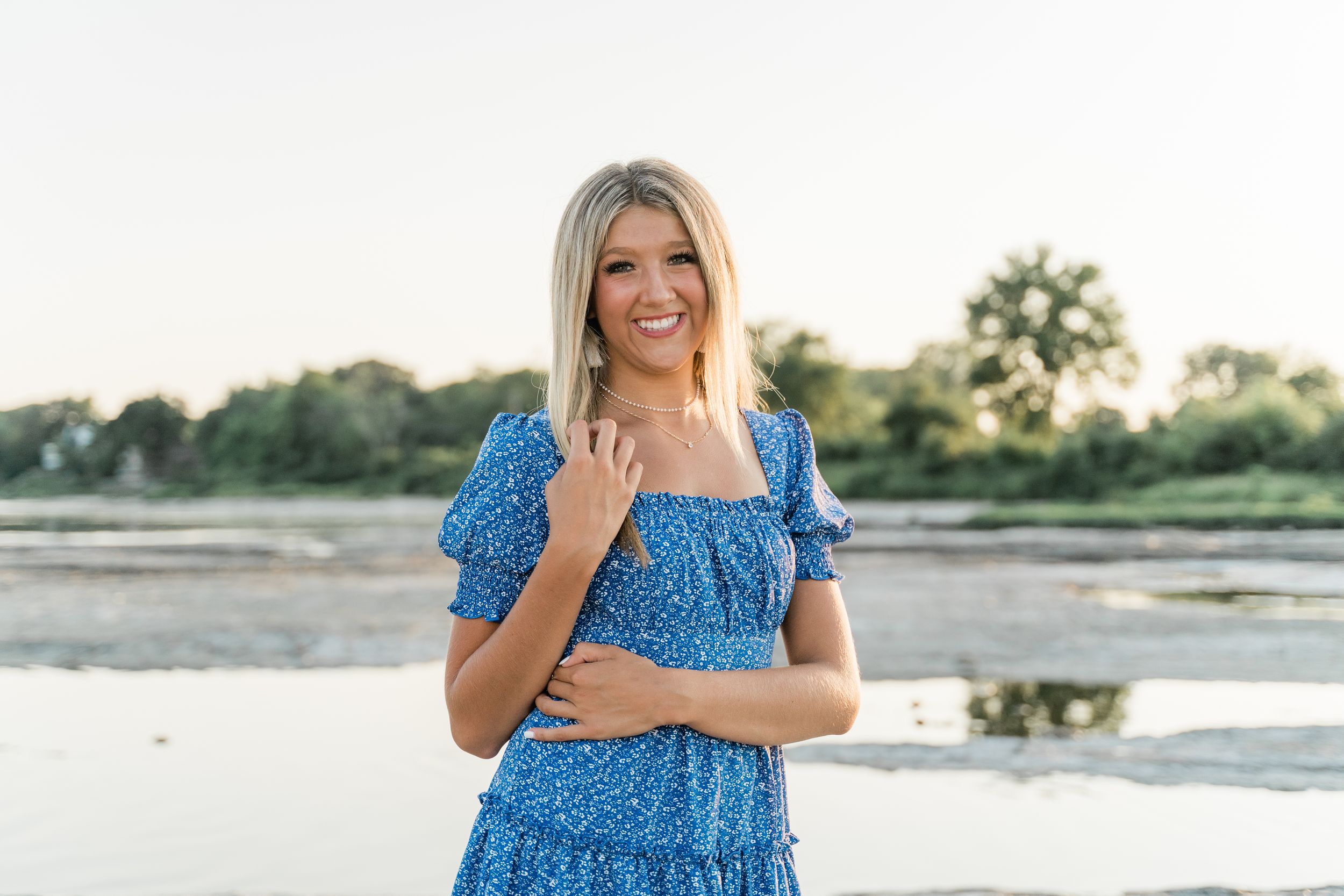 Woman in blue floral dress poses outdoors at sunset near a scenic waterfront.