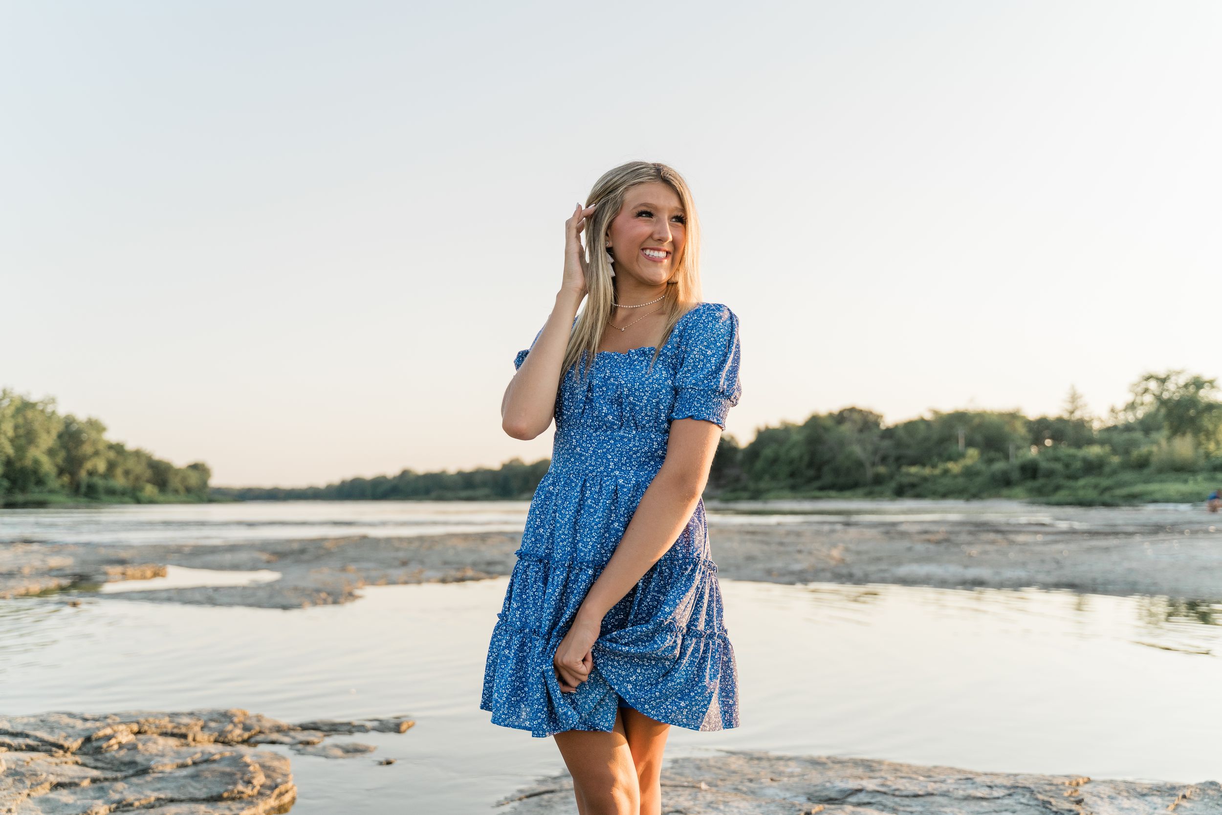 Woman wearing blue floral dress poses on sandy beach with water and trees in background.