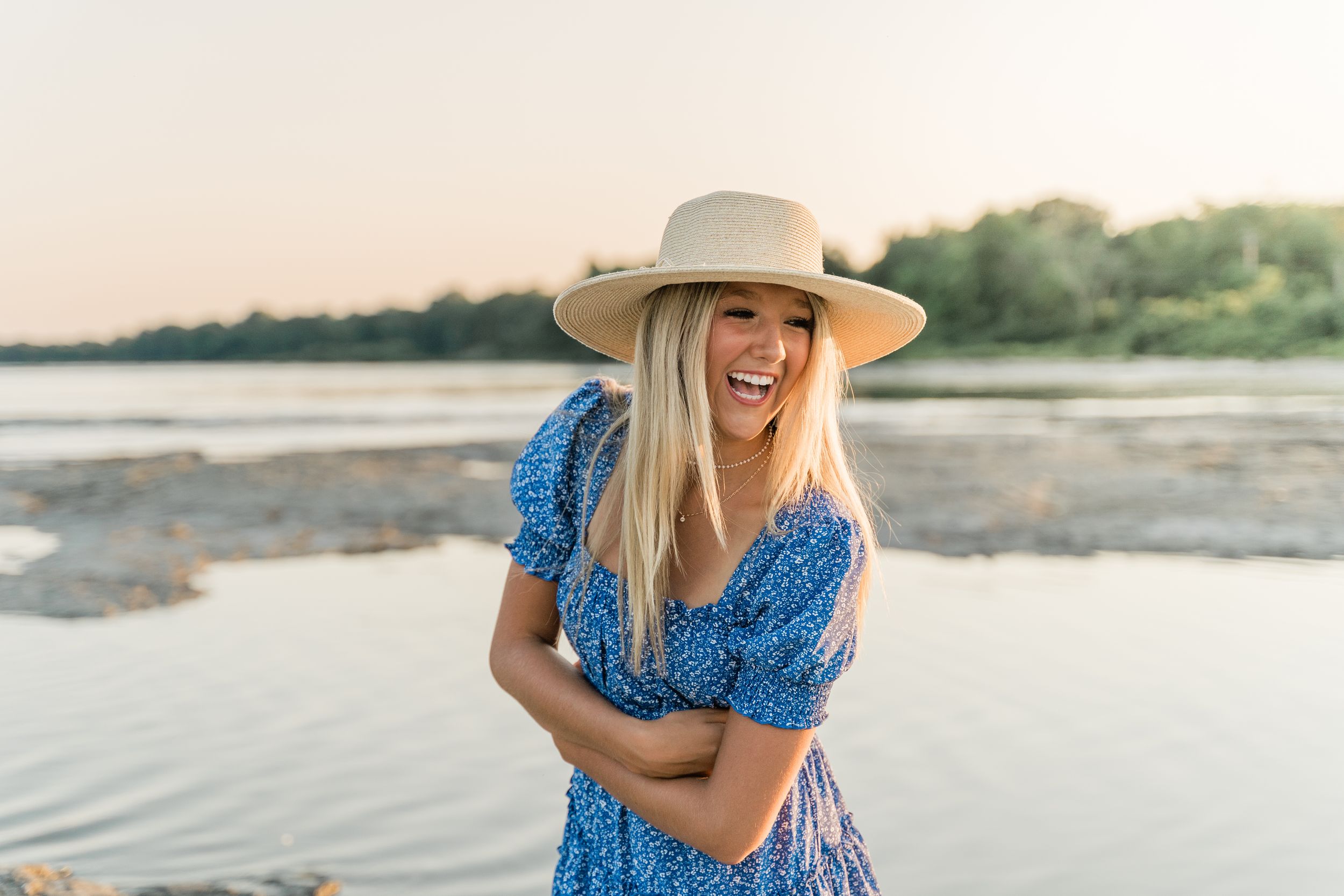 Woman in blue dress and straw hat enjoys sunset by the water's edge.