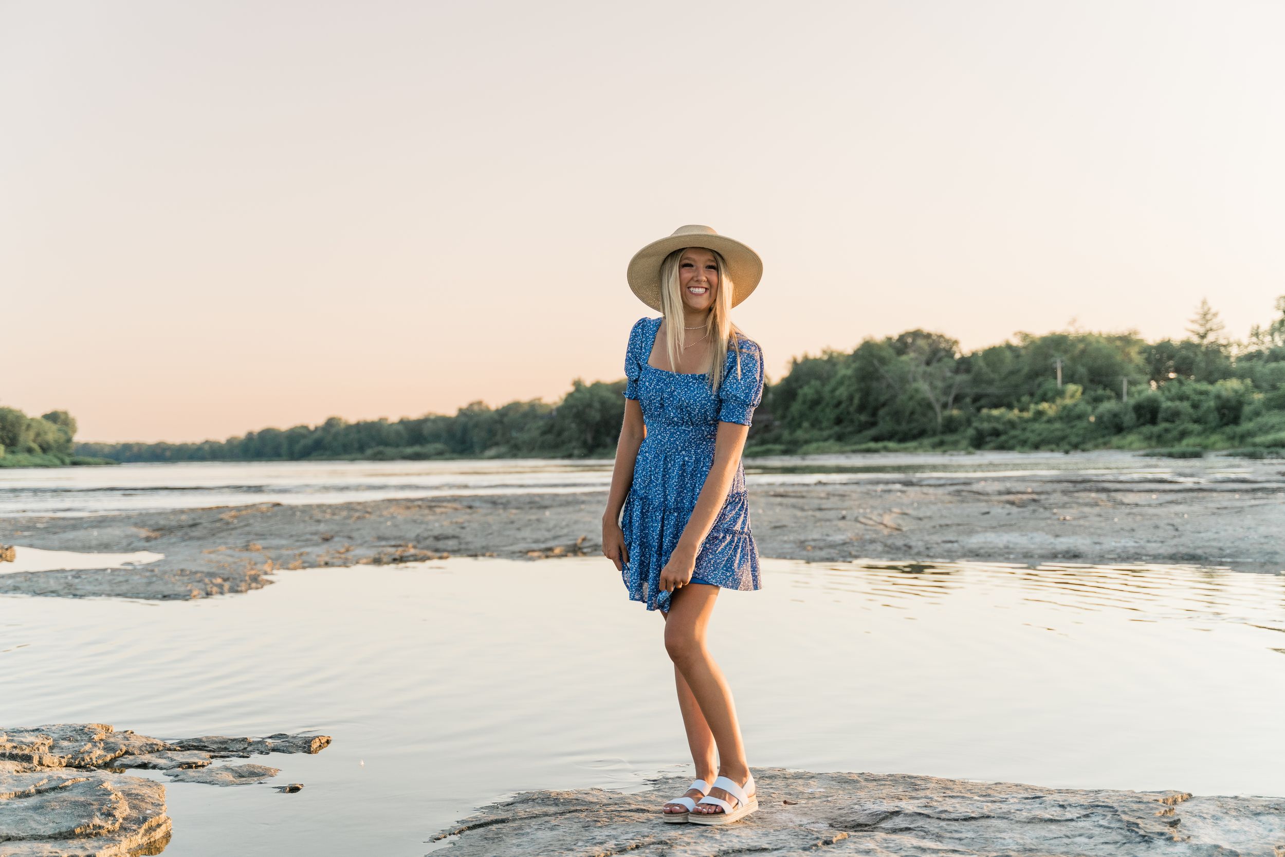 Someone in a blue sundress and sun hat stands on rocky terrain near a calm waterway during sunset.