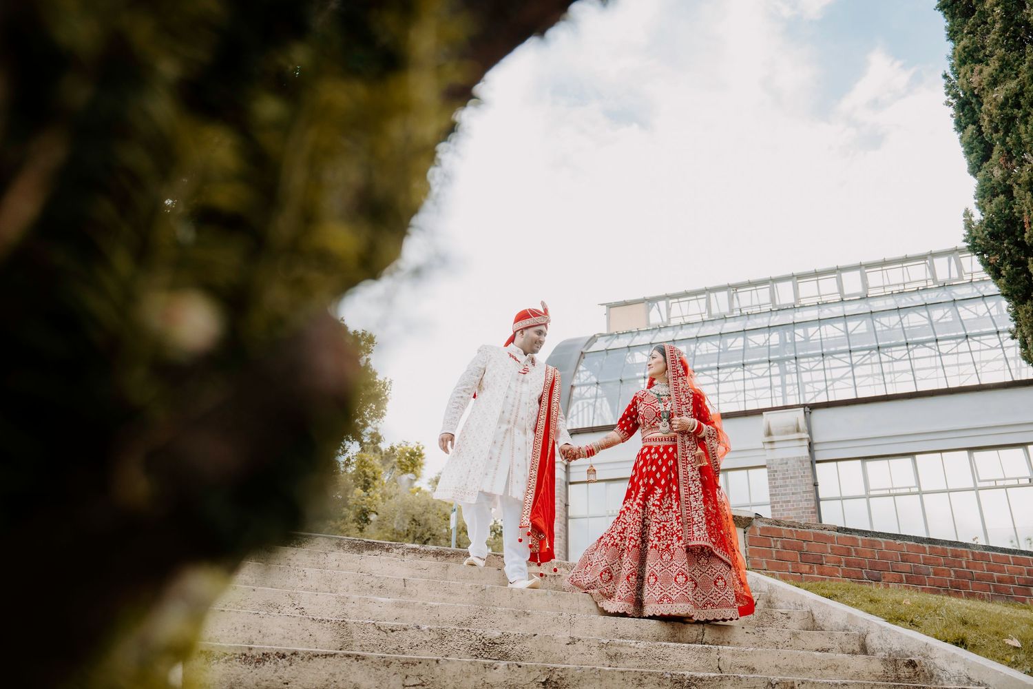A couple in traditional Indian wedding attire walks up stone steps at an outdoor venue during a wedding ceremony.