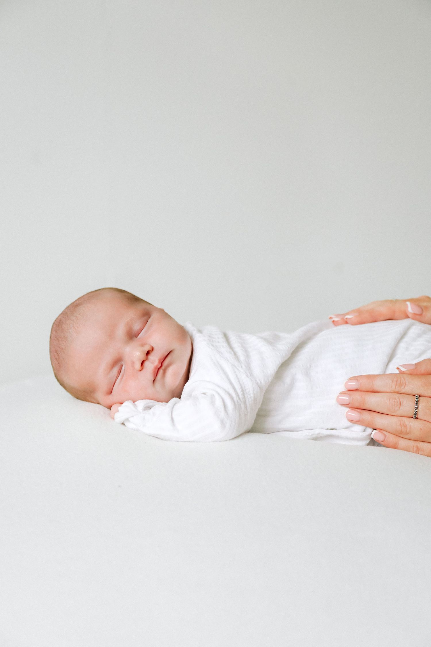 A sleeping newborn baby in white clothing rests peacefully as a gentle hand touches their side, during newborn shoot based in Nottingham