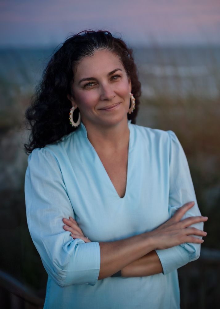 Woman in light blue top smiling warmly with arms crossed outdoors at dusk.